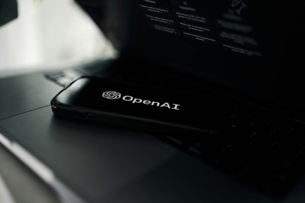 Móvil negro mostrando el logo y nombre de OpenAI apoyado sobre teclado de portátil en entorno oscuro